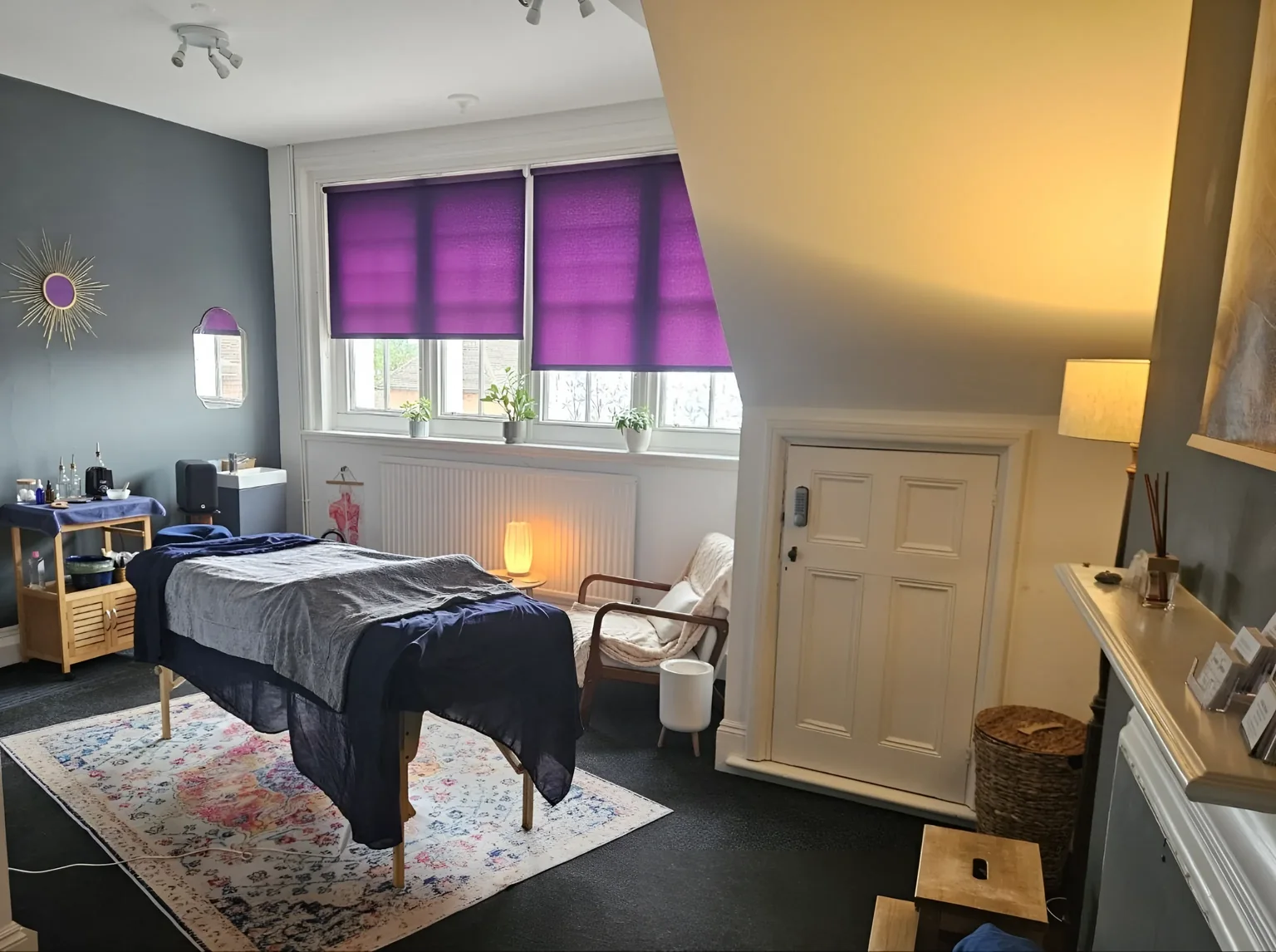 A treatment couch beneath purple window blinds in a bright massage room at The Treatment Rooms.